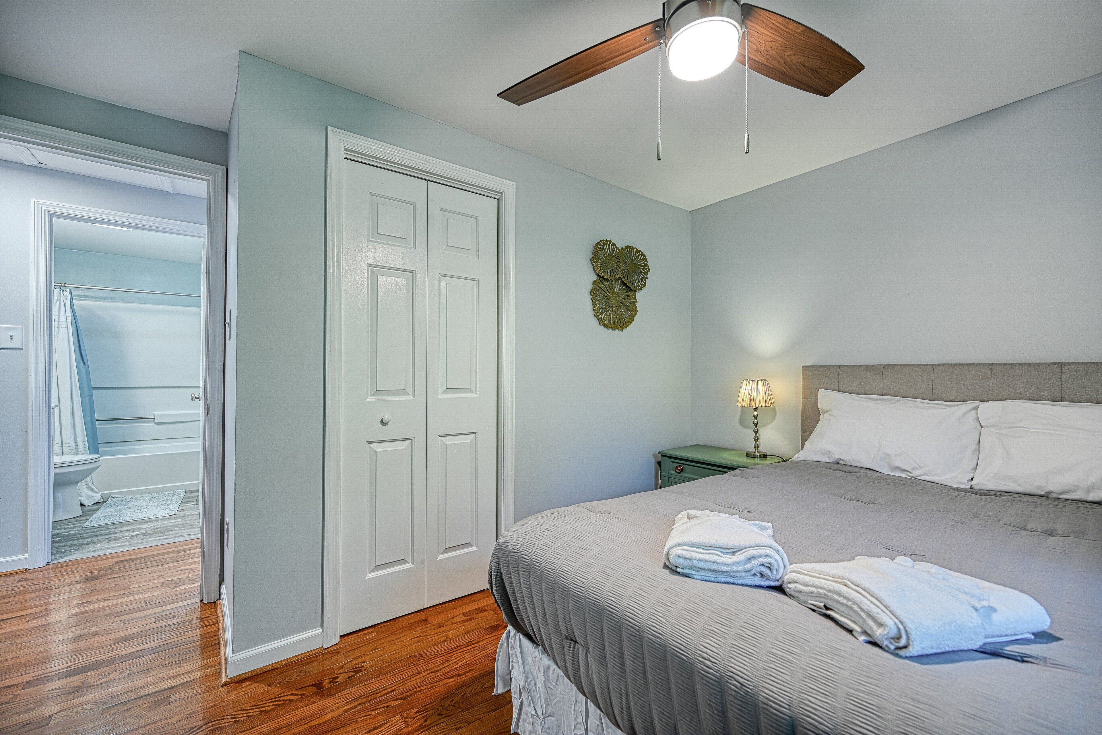 A bedroom with a bed, a ceiling fan, a nightstand with a lamp, and a door leading to a bathroom.