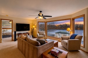 A living room with a sofa, chairs, a TV, and a view of a lake at sunset.