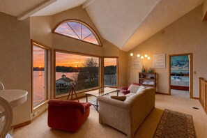 Uma sala de estar ampla com uma grande janela com vista para um lago ao pôr do sol, uma poltrona vermelha, um sofá bege e uma área de jantar.