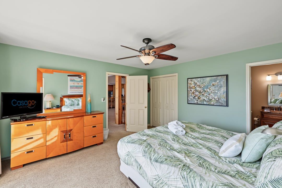 A bedroom with a bed, a television, a ceiling fan, and a door leading to another room.