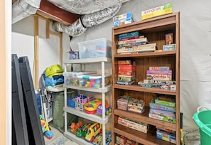 A room with a wooden bookshelf filled with board games, a white storage unit with toys, and a green bucket. - Lakefront Retreat With Views, hot Tub, Kid's Play Area, Dock & Fiber Internet (Mineral)