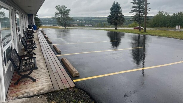 A covered outdoor area with a row of benches, a building with large windows, and a parking lot with yellow lines. - Ja-Sa-Le Motel (Bubartown)