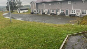 A row of single-story motel rooms with individual air conditioning units, a covered parking area, and a grassy landscape with a tree. - Ja-Sa-Le Motel (Bubartown)