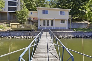 Eine Holzsteg, der zu einem Haus mit Balkon führt, umgeben von Grünpflanzen und einem Gewässer.