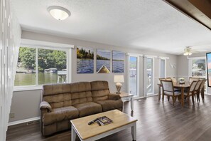 Ein Wohnzimmer mit Sofa, Couchtisch und Essbereich mit Stühlen. Große Fenster bieten Blick auf ein Gewässer und Boote.