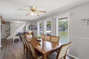 Ein Essbereich mit einem Holztisch und Stühlen, einem Deckenventilator und großen Fenstern mit Blick auf ein Gewässer.