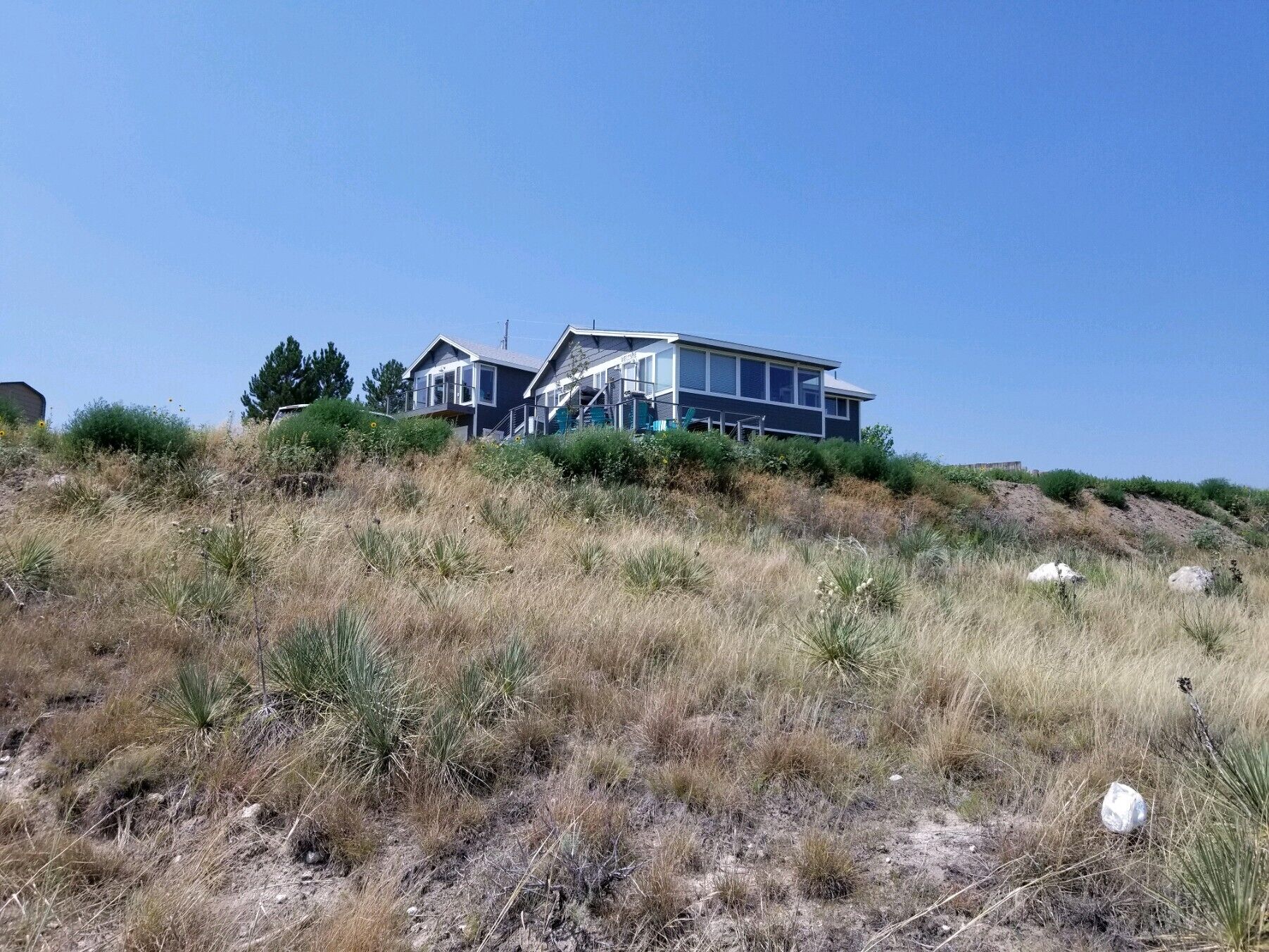 A modern two-story house with a flat roof and large windows, surrounded by dry grassland and a clear blue sky.