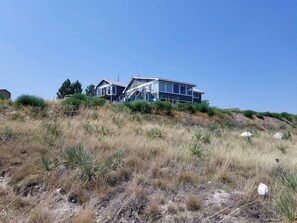 A modern two-story house with a flat roof and large windows, surrounded by dry grassland and a clear blue sky.