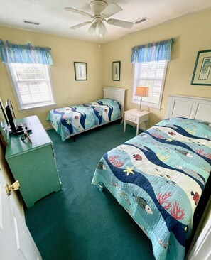 Une chambre avec deux lits, un bureau, une chaise, un ventilateur de plafond et deux fenêtres avec des rideaux.
