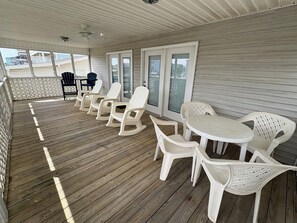 Un espace extérieur abrité, doté d’une terrasse en bois, de chaises en plastique blanches, d’une table ronde et d’un ventilateur de plafond.