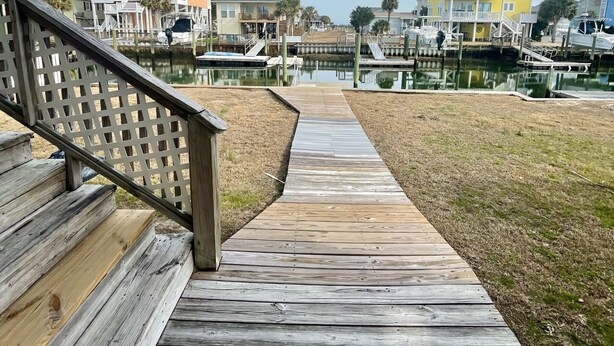 A wooden boardwalk leading to a marina with boats, a grassy area, and buildings with balconies. - Canal-front 3 Bed 2 Bath home including a dock. (Holden Beach)