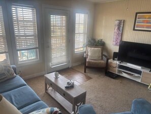 A living room with a sofa, coffee table, TV stand, and windows with blinds. - Canal-front 4BR, 2BA Holden Beach retreat with dock (Holden Beach)