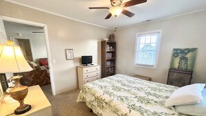 Ein Schlafzimmer mit einem Bett, Deckenventilator, Bücherregal, Schreibtisch mit Lampe und einem Fenster.