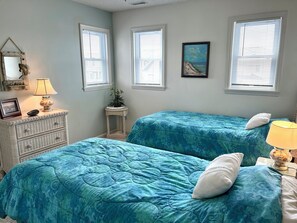 A bedroom with two beds, a dresser, a lamp, a framed picture, and a window with blinds. - Waterfront 4 bedroom retreat with dock and porch swing! (Holden Beach)