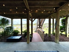 A covered outdoor area with a wooden deck, a staircase, and a view of a landscape with greenery and buildings. - Waterfront 4 bedroom retreat with dock and porch swing! (Holden Beach)