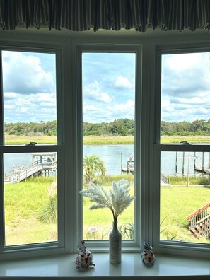 A window with a view of a lake, a dock, and a grassy area. - Waterfront 4 bedroom retreat with dock and porch swing! (Holden Beach)