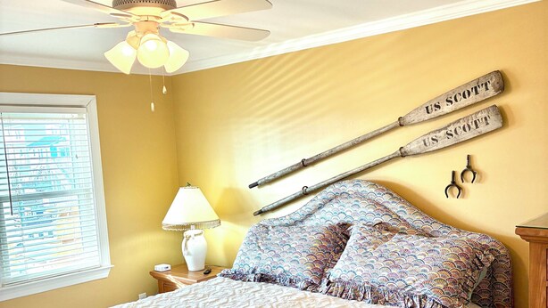A bedroom with a bed, a ceiling fan, a window with blinds, a bedside table with a lamp, and a wall-mounted baseball bat. - Spacious 3-bedroom, 2-bath canal home located with Canal access and Waterway views. (Supply)
