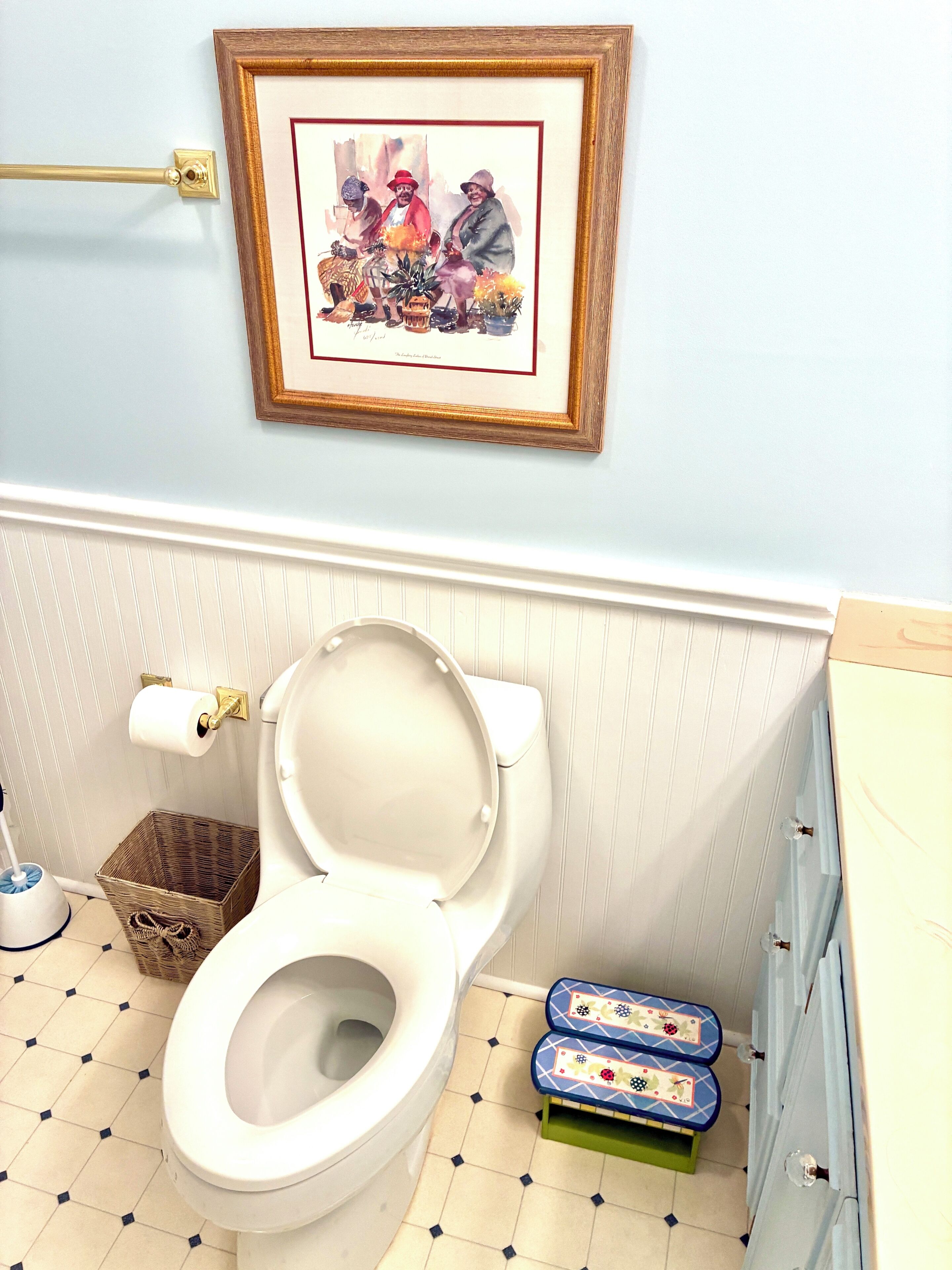A bathroom with a toilet, a framed picture on the wall, a basket, and a small stool.