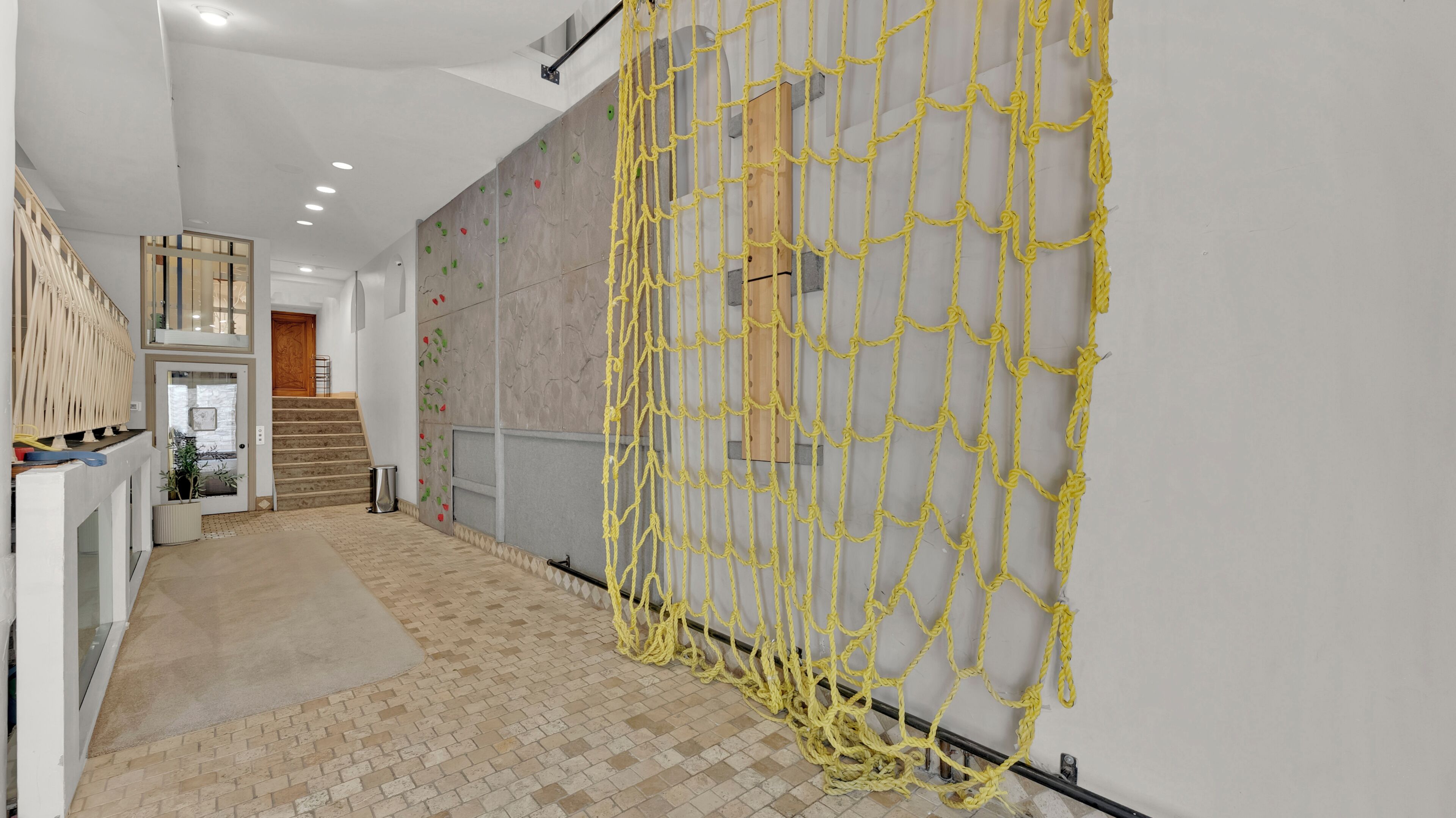 Un couloir avec un mur d’escalade équipé de prises jaunes, un escalier et une porte donnant sur une autre pièce.