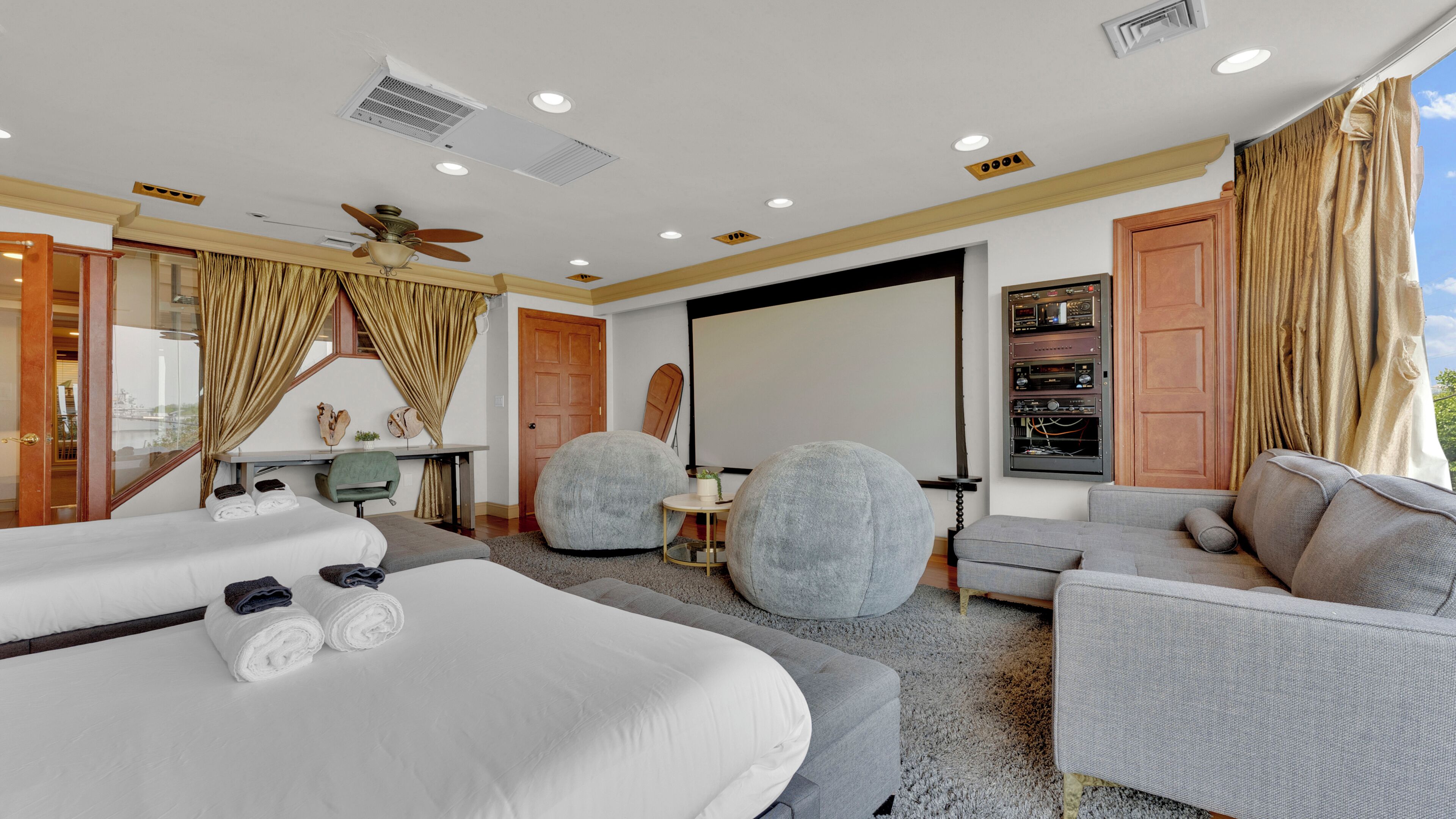 A hotel room with a bed, two grey sofas, a small table, a ceiling fan, and a projector screen.