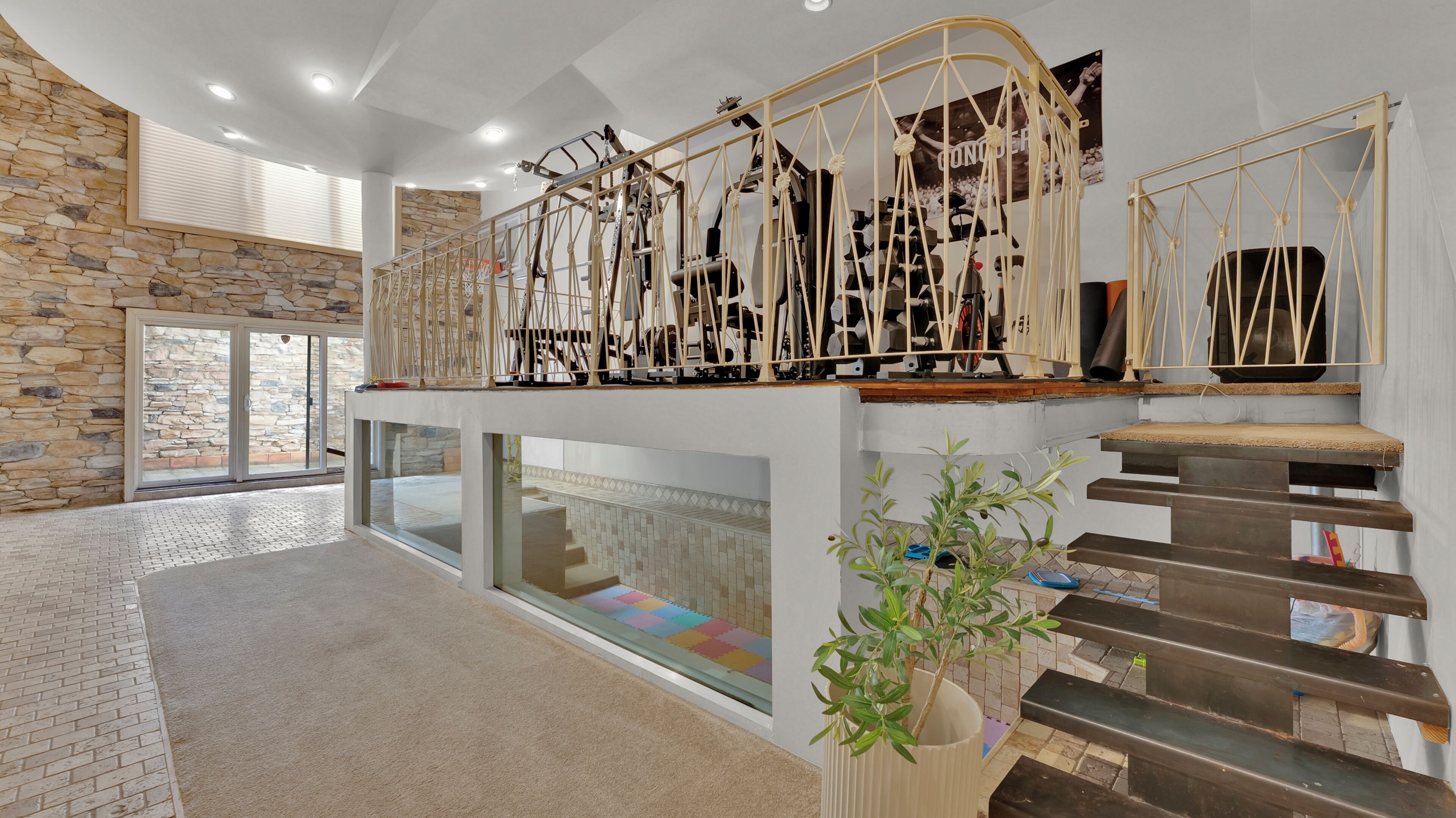 A spacious interior with a stone wall, a glass door leading outside, a staircase with a railing, and a large potted plant.