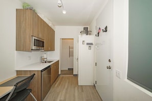 A compact kitchen with wooden cabinets, a microwave, a refrigerator, and a small dining area with a chair.