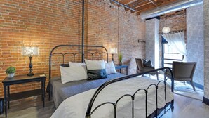 A room with a brick wall, a metal bed frame, a small table with a lamp, and a chair.