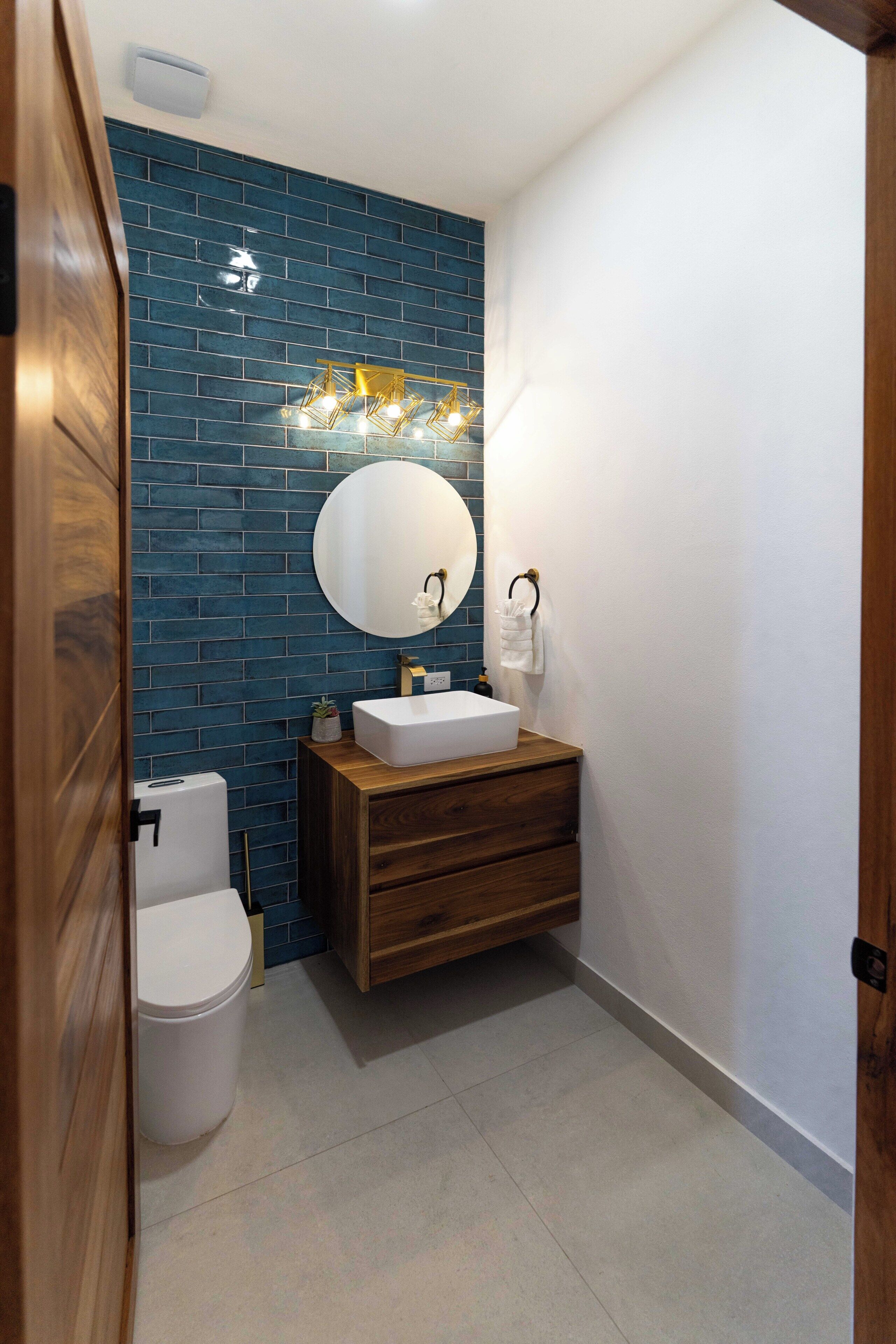 A bathroom with a blue-tiled wall, a wooden vanity, a white sink, and a round mirror.