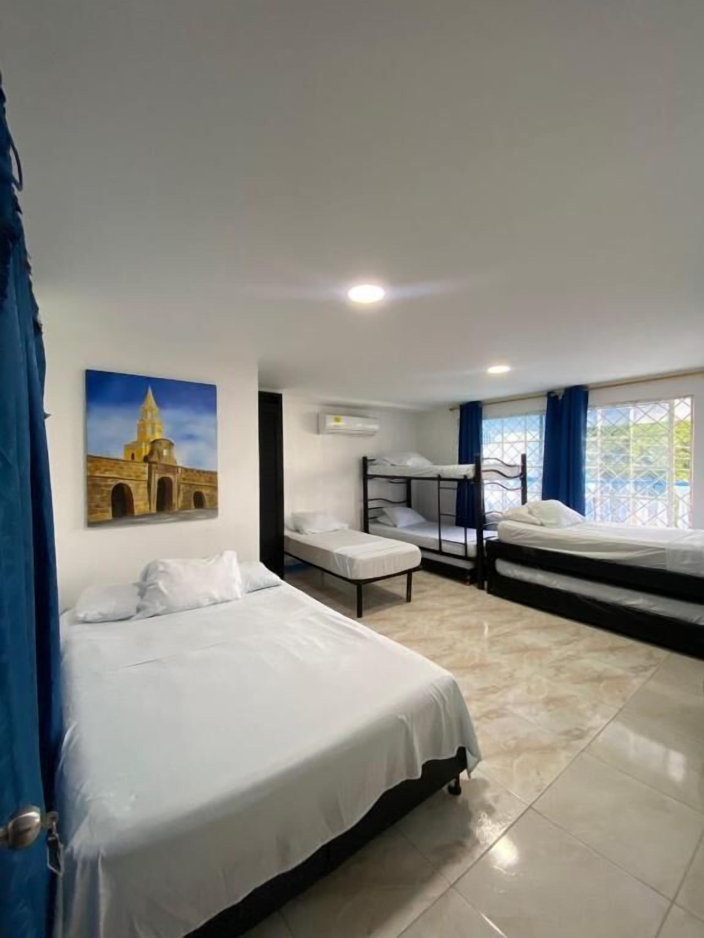 A hotel room with two beds, a bunk bed, a window with blue curtains, and a wall-mounted air conditioner.
