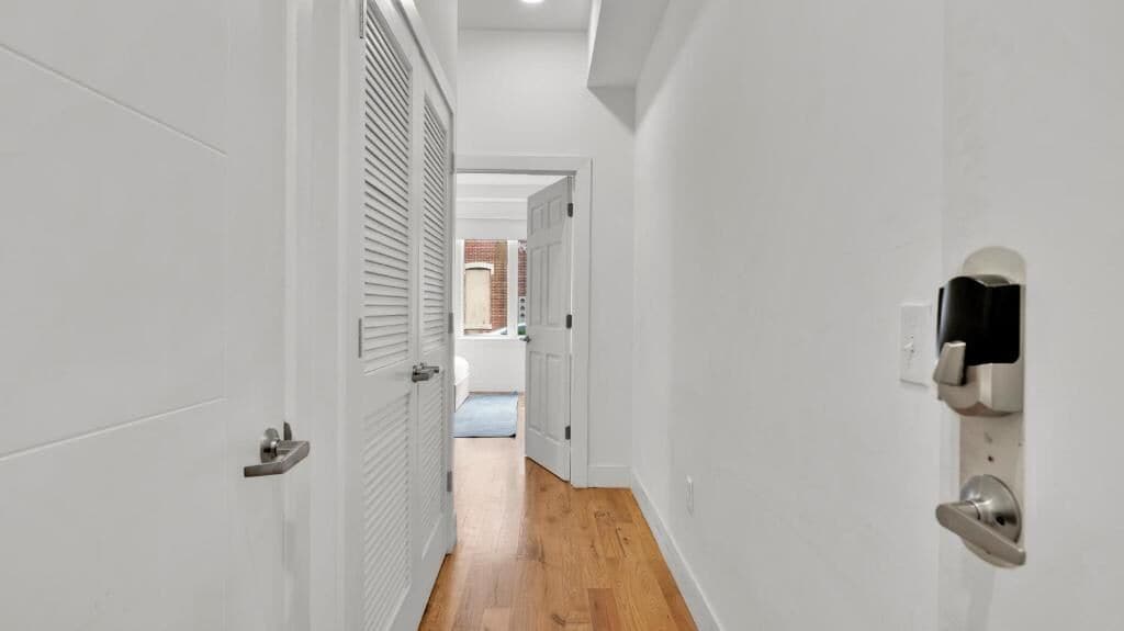 A narrow hallway with white walls and wooden flooring, leading to a room with a door and a window.