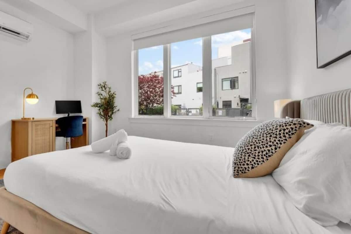 Une chambre d’hôtel moderne avec un grand lit, un bureau avec une lampe, une télévision et une fenêtre donnant sur des immeubles.