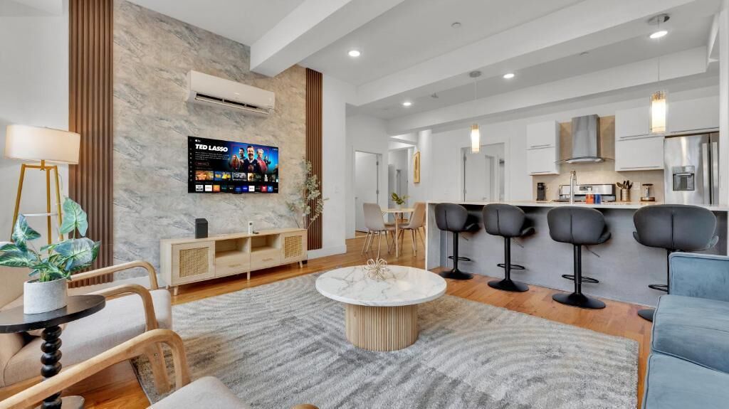 Een moderne woonkamer met een marmeren salontafel, een aan de muur bevestigde televisie en een keuken met barstoelen.