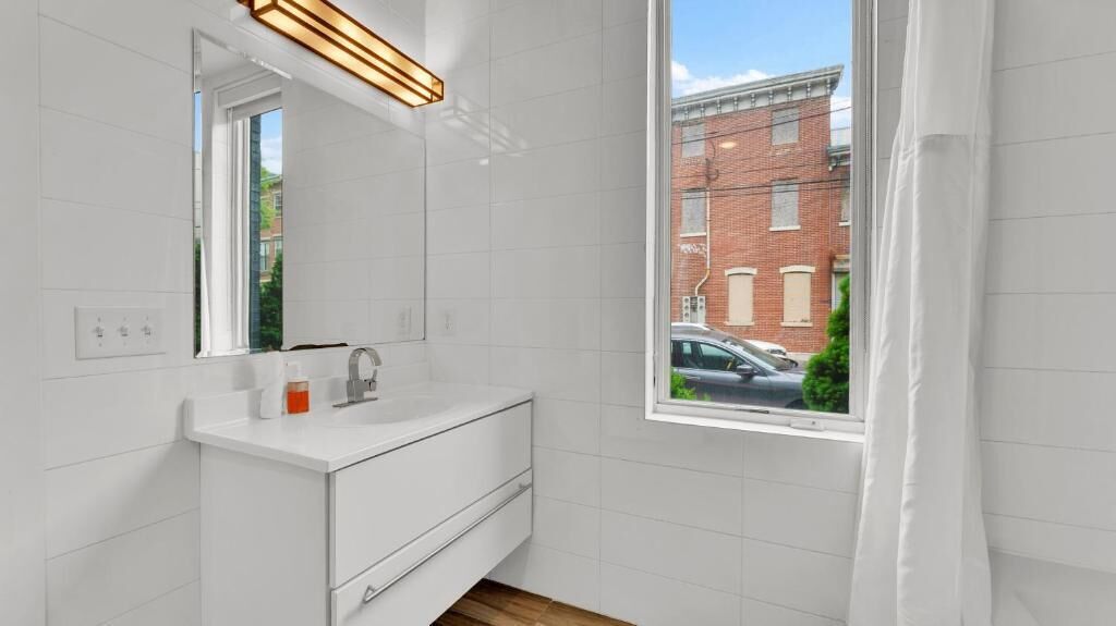 Une salle de bain moderne avec des carreaux blancs, un grand miroir, un plan de toilette blanc et une fenêtre donnant sur des immeubles.