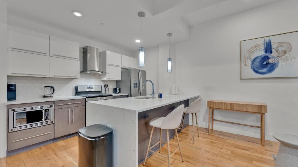 A modern kitchen with white cabinets, stainless steel appliances, a central island with bar stools, and a wooden table.