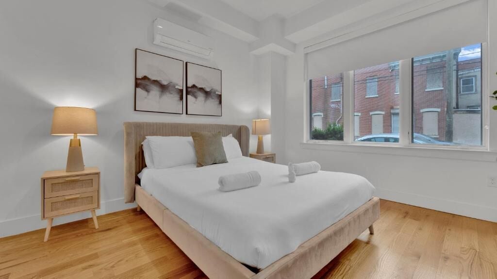Ein Schlafzimmer mit einem Bett, zwei Nachttischen, einem Fenster mit Blick auf Gebäude und Kunstwerken an der Wand.
