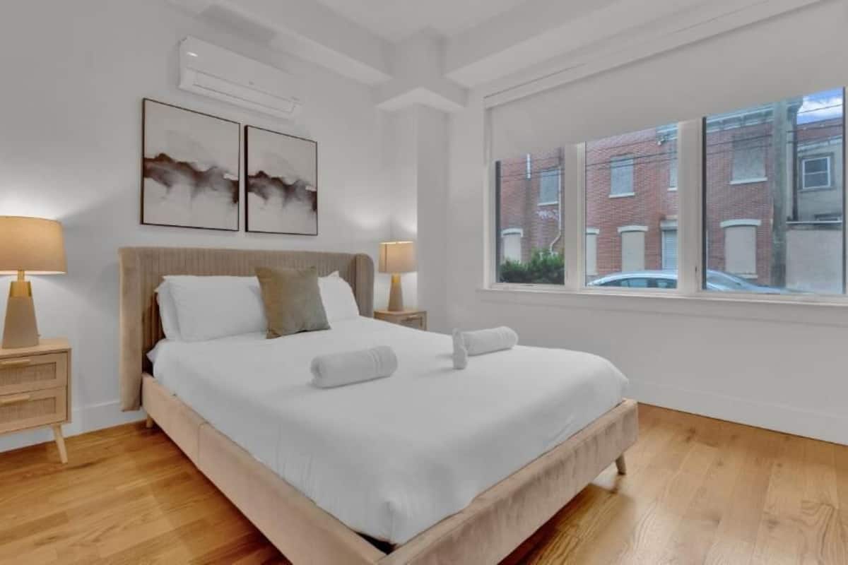 Une chambre à coucher avec un lit, deux tables de chevet, une fenêtre donnant sur des immeubles et des œuvres d’art accrochées au mur.