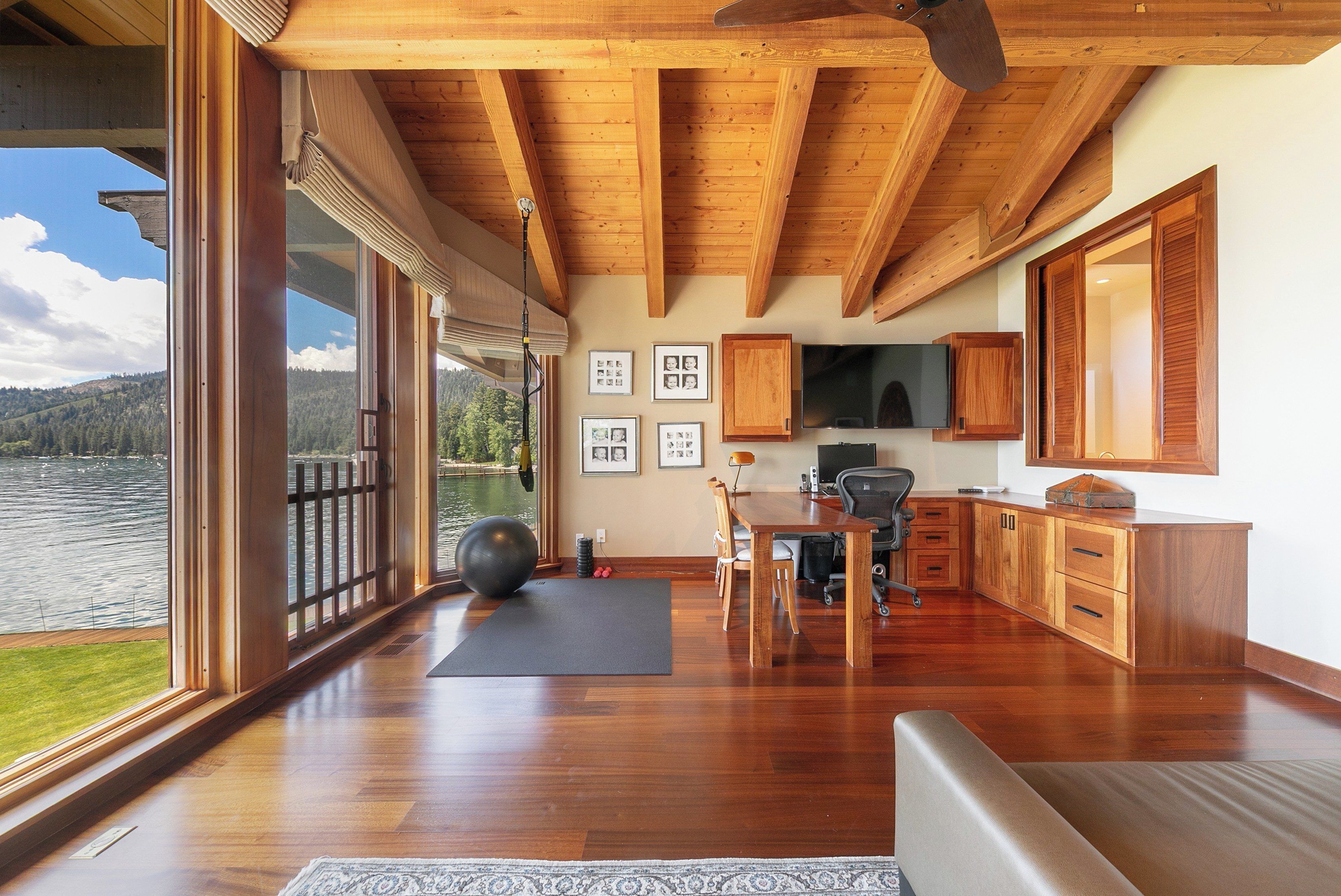 A spacious room with a large window overlooking a body of water, wooden flooring, a desk with a chair, and a television mounted on the wall.