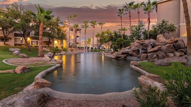 Una piscina di un resort circondata da palme e rocce, con un tramonto sullo sfondo.
