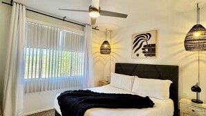 Una camera da letto con un letto, una ventola a soffitto, una finestra con tende, una lampada da comodino e un'opera d'arte appesa al muro.