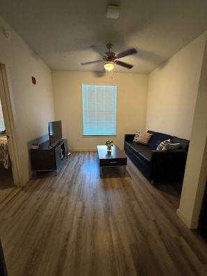 Una sala de estar con ventilador de techo, televisor sobre un mueble bajo, una mesa de centro con una planta, sofá con cojines estampados y una ventana con persianas.
