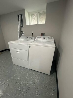 Een wasserette met een voorlader wasmachine en droger, een raam boven de droger, en een douchestang.