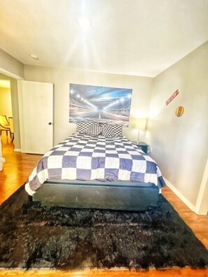 Een slaapkamer met een geruit beddegoed, een pluizig zwart vloerkleed, een aan de muur hangend kunstwerk en een deur naar een andere kamer.