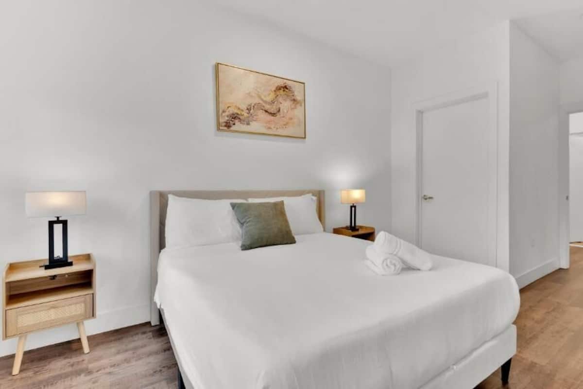 Une chambre à coucher comprenant un lit, des tables de chevet, une lampe, un tableau et une porte donnant sur une autre pièce.