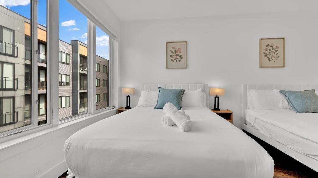 Hotellihuone, jossa on suuri sänky, kaksi yöpöytää, ikkuna, josta on näkymä rakennuksiin, ja kaksi seinälle kiinnitettyä valokuvakehystä.