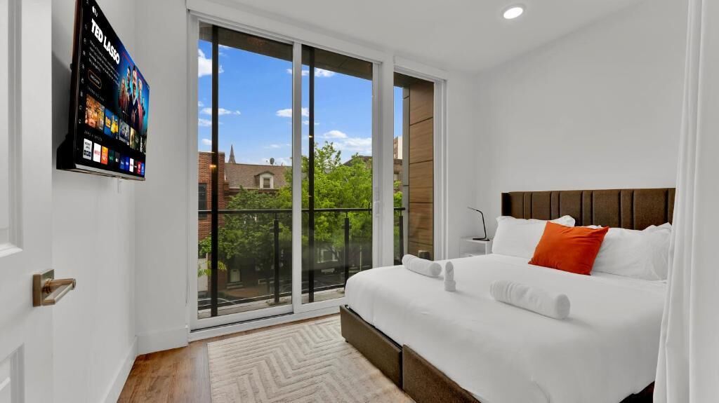 A modern hotel room with a large bed, a TV mounted on the wall, and a sliding glass door leading to a balcony.