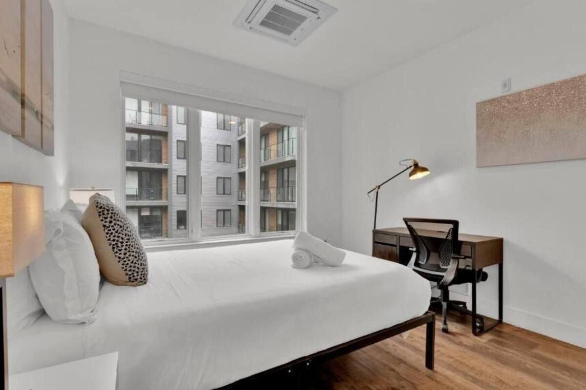 Une chambre à coucher avec un lit, un bureau, une chaise, une lampe et une vue sur un immeuble par la fenêtre.