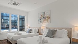 A hotel room with two beds, a large window, a painting on the wall, and a bedside table with a lamp. - Apartmint at Liberty A Philadelphia (Philadelphia)