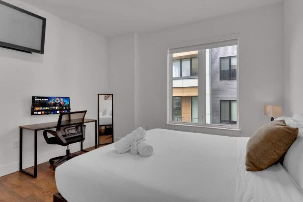 Une chambre d’hôtel avec un lit, un bureau et une chaise, une télévision et une fenêtre donnant sur des immeubles.