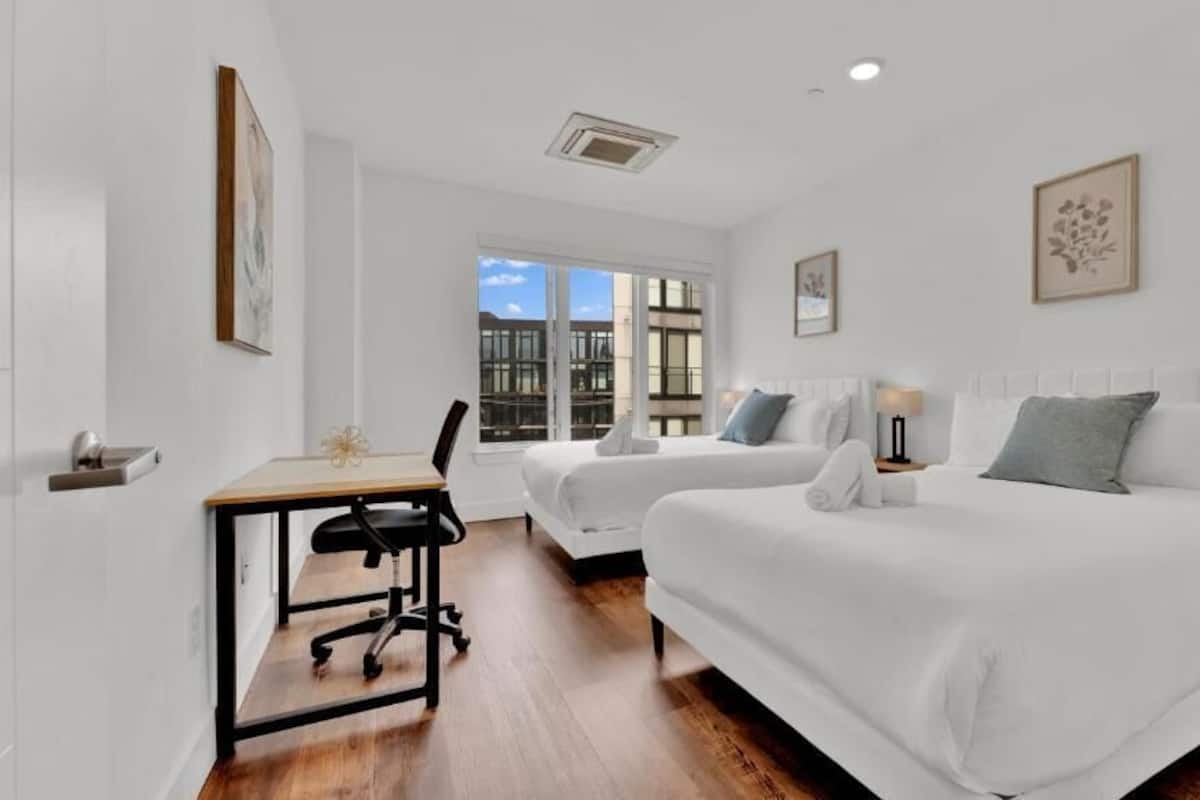 Une chambre d’hôtel avec deux lits, un bureau, une chaise et une fenêtre donnant sur des immeubles.