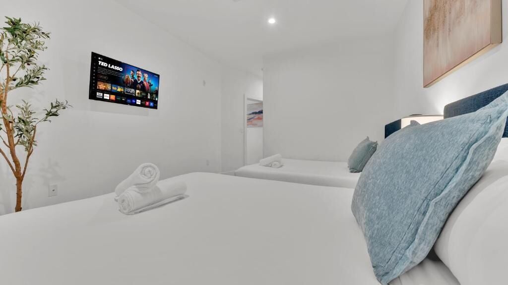 A modern hotel room with a flat-screen TV, two beds, a plant, and a painting on the wall.